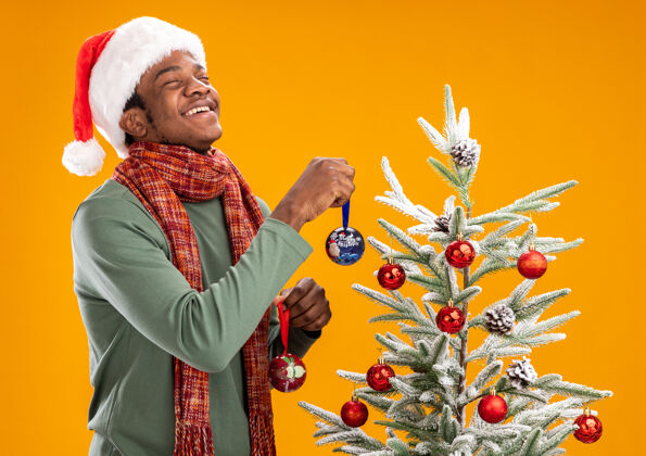 帽子戴着圣诞帽 围着围巾的非洲裔美国人把圣诞球挂在圣诞树上快乐地站在橙色的墙上非洲周围站