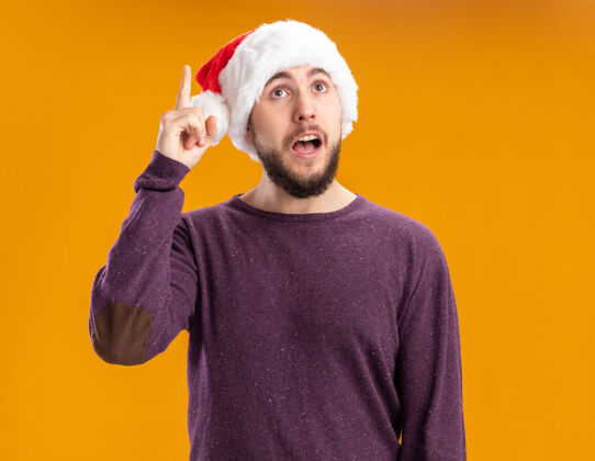 显示一个穿着紫色毛衣 戴着圣诞帽的年轻人惊讶地抬起头来 站在橙色的墙上 伸出食指 有了新的好主意圣诞节帽子站立