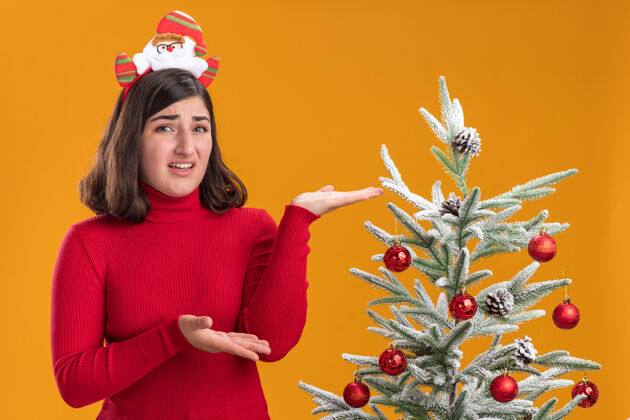 圣诞节穿着圣诞毛衣的年轻女孩戴着有趣的头带 站在橙色背景下的圣诞树旁毛衣礼物圣诞树