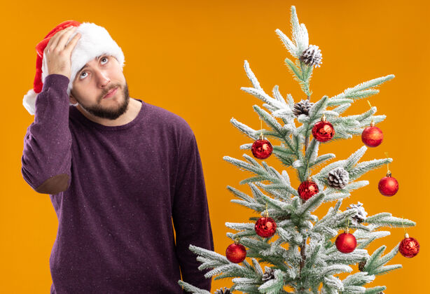 年轻一个穿着紫色毛衣 戴着圣诞帽的年轻人站在橙色背景下的圣诞树旁 看上去很困惑 很焦虑毛衣男人困惑