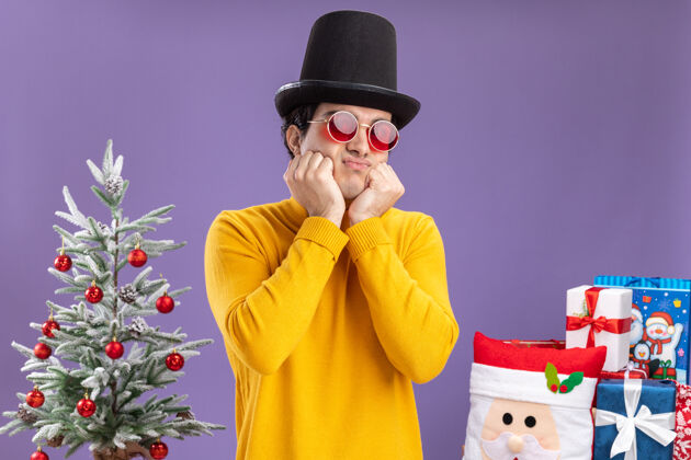 礼物一个穿着黄色高领毛衣 戴着黑帽子的悲伤的年轻人站在圣诞树旁 在紫色背景下送礼物帽子站着穿着