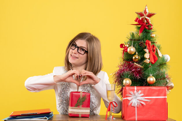 礼物正面图：女医生坐在桌子前 拿着礼物 黄色背景上的圣诞树和礼盒成人微笑开朗