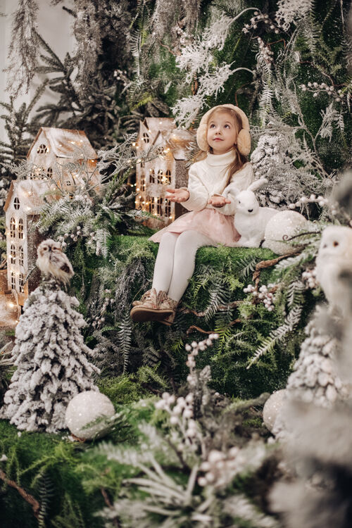 魔术美丽的白种人小孩 长着金色的长发 微笑着坐在圣诞气氛中 周围有很多装饰树 还有一只小兔子自然庆祝新