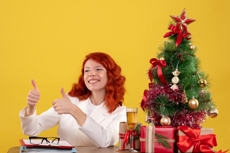 桌子女工人坐在桌子后面 拿着圣诞树和黄色的礼物黄色女圣诞节