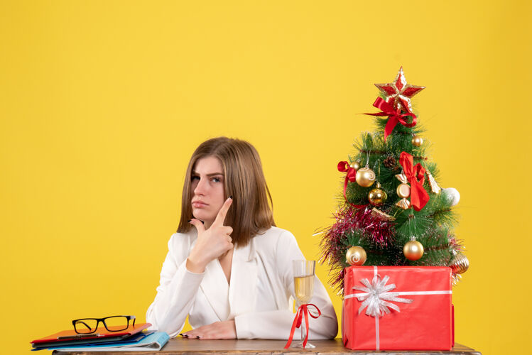 情感正面图：女医生坐在黄色背景的桌子前 放着圣诞树和礼品盒办公室健康女性