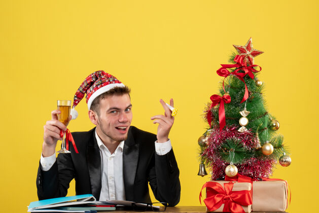 香槟正面图：男员工在小圣诞树周围用香槟庆祝圣诞节 黄色办公桌上的礼物新年办公室颜色圣诞节颜色男性周围