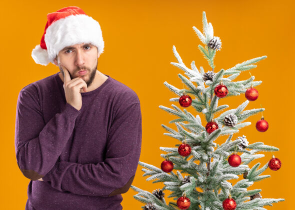 圣诞树身穿紫色毛衣 戴着圣诞帽的年轻人在橙色背景下的圣诞树旁沉思地看着摄像机毛衣年轻人帽子