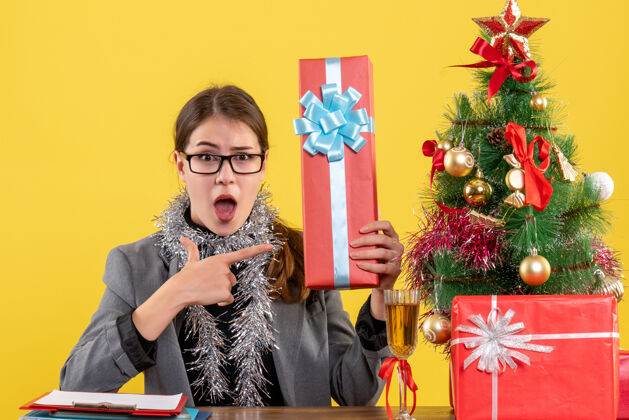 人正面图：戴眼镜的大眼睛女孩坐在桌旁 展示圣诞树礼物和鸡尾酒礼物展示礼物女孩
