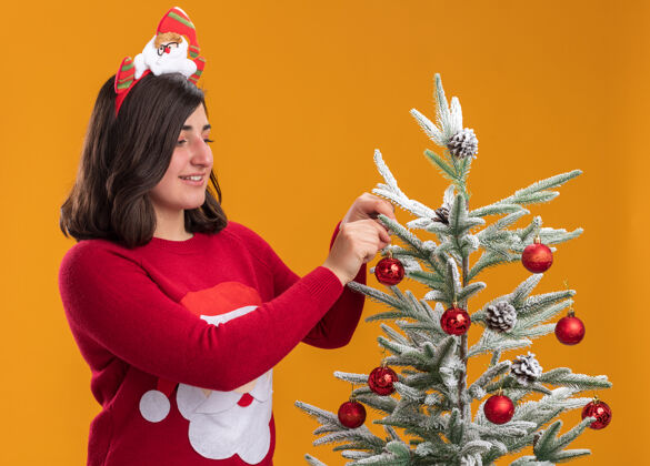 穿穿着圣诞毛衣 戴着滑稽头带的快乐小女孩站在圣诞树旁 圣诞树上挂着圣诞球 挂在橙色的墙上毛衣站挂