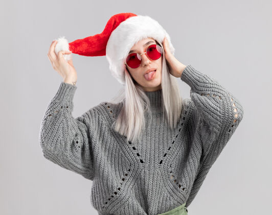 粘快乐的年轻金发女人穿着冬季毛衣 戴着圣诞帽 看起来很开心伸出舌头快乐舌头快乐
