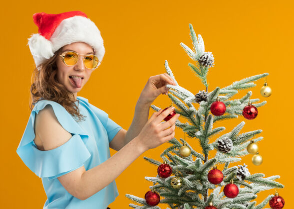 圣诞身着蓝色上衣 戴着圣诞帽 戴着黄色眼镜 站在橙色背景下 装饰圣诞树 伸出舌头的快乐快乐的年轻女子站立眼镜年轻