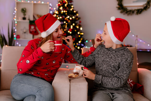 装饰快乐的妈妈和小儿子戴着圣诞帽 喝着茶 坐在沙发上吃着饼干 在装饰好的房间里玩得很开心 背景是圣诞树圣诞老人房间坐