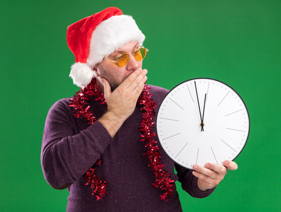 中年一个戴着圣诞老人帽 脖子上戴着金属丝花环 戴着眼镜 手放在嘴上 手举着手 看着隔离在绿色墙上的钟的关心的中年男人关心时钟保管
