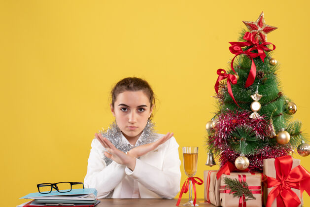 坐着正面图：女医生坐在黄色背景的桌子后面 还有圣诞树和礼品盒快乐背景人