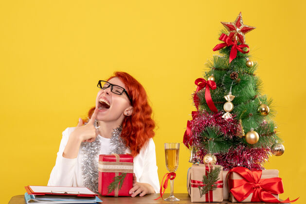 圣诞节前视图：女医生坐在桌子后面 黄色背景上有圣诞礼物前面插花女医生