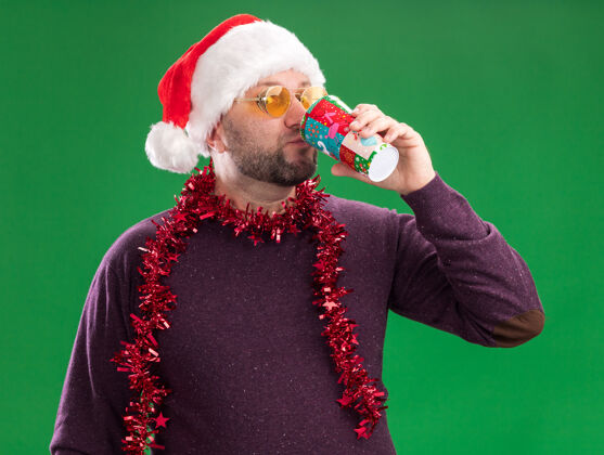 喝酒中年男子戴着圣诞帽 脖子上戴着金属丝花环 戴着眼镜看着相机喝着咖啡周围帽子中年