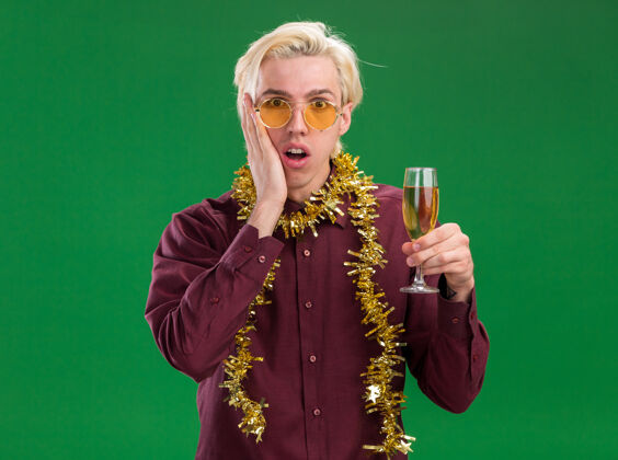 穿着关心的年轻金发男子戴着眼镜 脖子上戴着金箔花环 手里拿着香槟酒杯 手放在脸上 隔离在绿色的墙上香槟年轻脖子