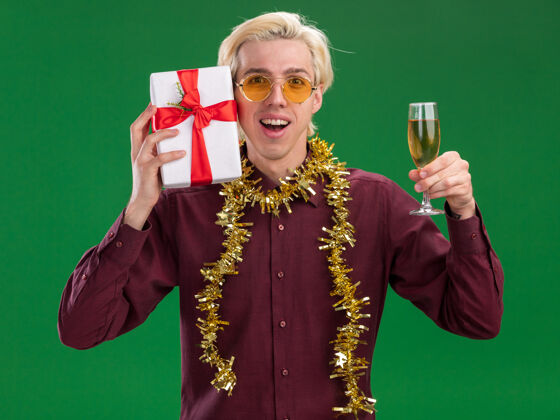 周围令人印象深刻的年轻金发男子戴着眼镜 脖子上戴着金箔花环 手持香槟酒杯 用隔离在绿色墙上的礼包摸着头圣诞节香槟年轻