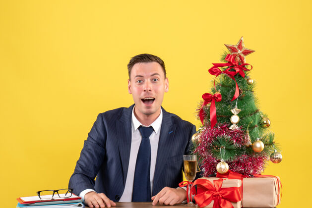 快乐男人圣诞树和黄色礼物旁的快乐男人坐在桌子前的正面视图商人快乐男人