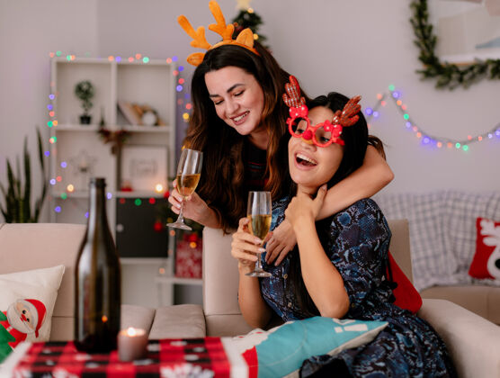 扶手椅戴着驯鹿头带的年轻女孩微笑着 手里拿着一杯香槟 抱着坐在扶手椅上的戴着驯鹿眼镜的朋友 在家里享受圣诞节时光圣诞节微笑时间