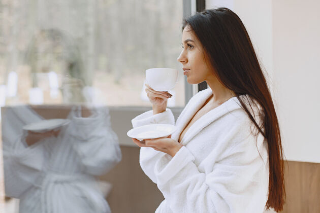 房间早上好女人喝咖啡靠窗的女人护理浴袍早晨