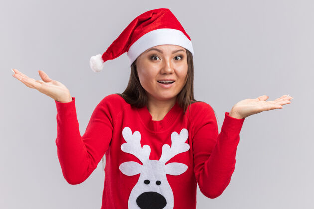 毛衣惊讶的年轻亚洲女孩戴着圣诞帽和毛衣摊开双手隔离在白色的墙上圣诞节女人惊喜