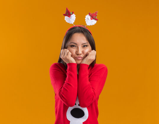 高兴高兴的年轻亚洲女孩戴着圣诞发箍和毛衣把拳头放在脸颊上隔离在橙色的墙上脸颊年轻拳头