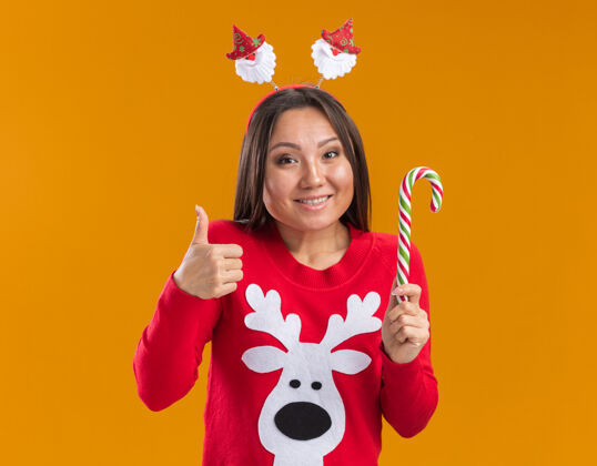 年轻笑容满面的年轻亚洲女孩 戴着圣诞发箍 穿着毛衣 拿着圣诞糖果 在橙色的墙上孤立地竖起大拇指穿女人拇指