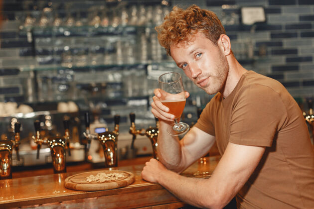 西装有魅力的年轻人站在吧台后面一个穿着棕色t恤的男人手里拿着一个玻璃杯成人啤酒顾客