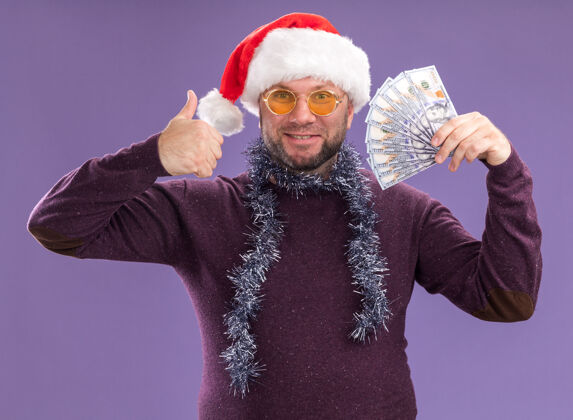 中年微笑的中年男子戴着圣诞帽 脖子上戴着金箔花环 戴着眼镜 手里拿着钱 在紫色的墙上孤立地竖起大拇指帽子圣诞节男人
