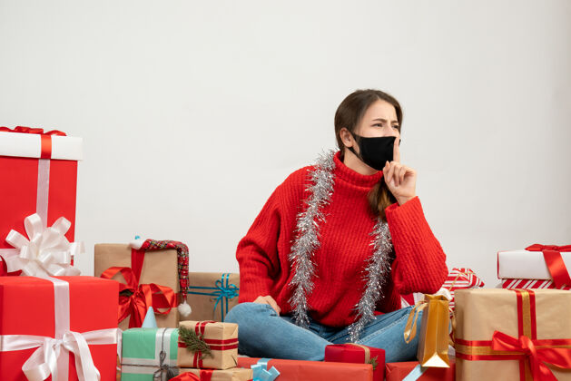 礼物穿着红色毛衣 戴着黑色面具的年轻女孩在白色的礼物旁做嘘手势圣诞节购物周围