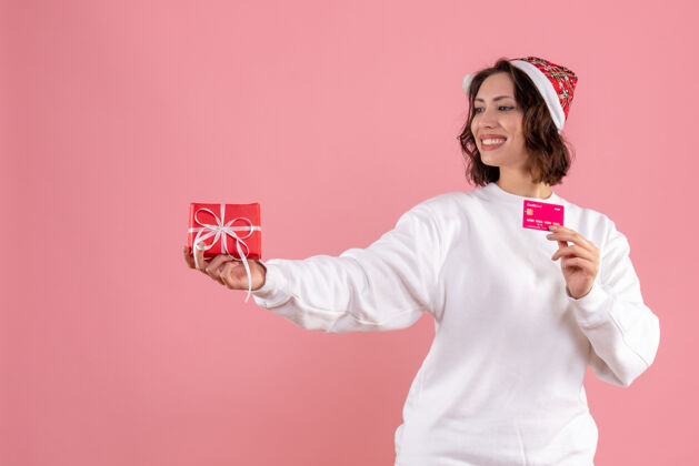 年轻的女性年轻女子手持小圣诞礼物和粉色墙上的银行卡的正视图人圣诞节情感