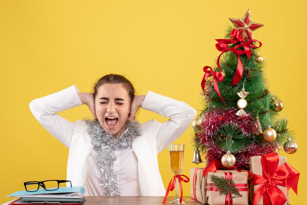 背景正面图：女医生坐在桌子后面 用圣诞树和礼盒把耳朵贴在黄色背景上关门圣诞节人
