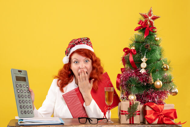 成人正面图：女医生坐在桌子后面 拿着黄色背景的计算器 还有圣诞树和礼品盒计算器桌子坐着