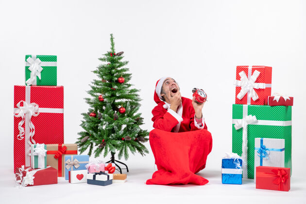 地圣诞老人坐在地上看上面 在礼物和圣诞树旁展示钟表礼物装饰人