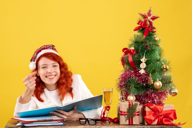 女性正面图：女医生坐在桌子后面看黄色背景上的文件 还有圣诞树和礼品盒礼物圣诞节健康