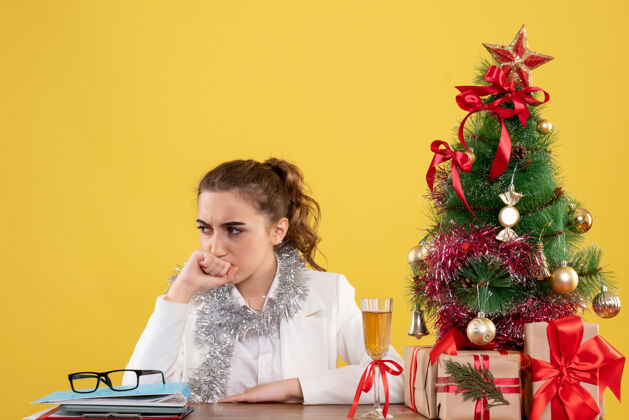 紧张正面图：女医生坐在桌子后面 脸上带着紧张的表情 背景是黄色的圣诞树和礼盒护士女医生前面