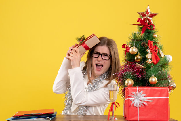 背景正面图：女医生坐在桌子前 手里拿着黄色背景的礼物 还有圣诞树和礼品盒健康办公室桌子