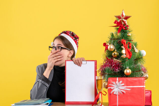 好奇前视图好奇的女孩戴着圣诞帽坐在桌旁 手里拿着文件夹圣诞树和鸡尾酒礼物树人桌子