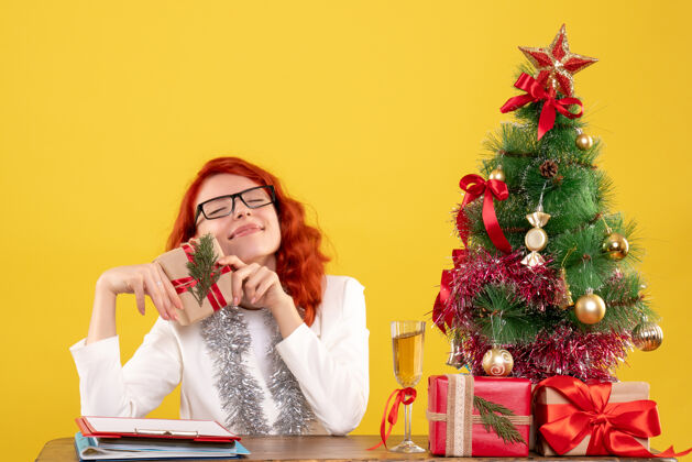 情感前视图：女医生坐在桌子后面 黄色背景上有圣诞礼物 圣诞树和礼品盒护士快乐肖像
