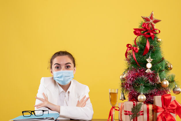 情感正面图女医生戴着防护面具坐在黄色背景上 戴着圣诞树和礼品盒女性病毒医疗