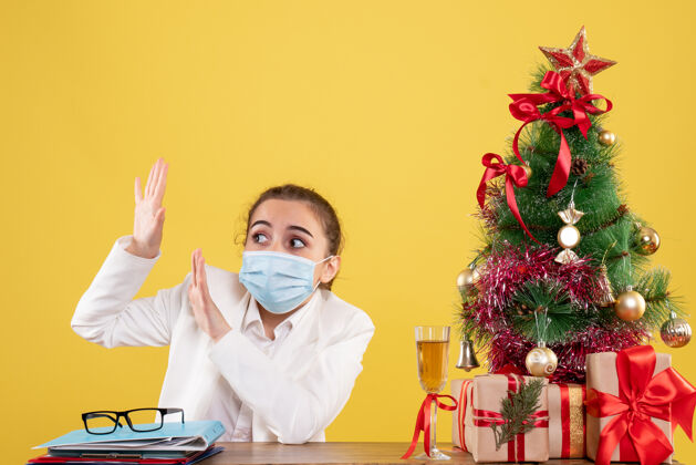 坐着正面图女医生戴着无菌口罩坐在黄色背景上 带着圣诞树和礼品盒男性专业前面