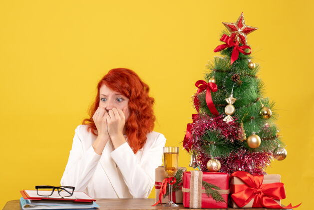 新的前视图：女医生坐在桌子后面 黄色背景上有圣诞礼物医生情感微笑