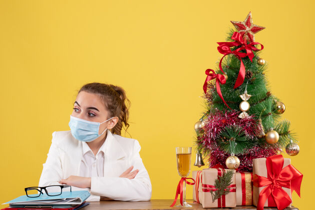 病毒正面图女医生戴着防护面具坐在黄色背景上 带着圣诞树和礼品盒背景人成人