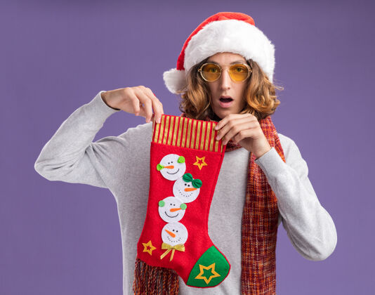 快乐年轻人戴着圣诞老人帽 戴着黄色眼镜 脖子上围着暖和的围巾 手里拿着圣诞长袜 站在紫色背景下惊讶地看着镜头圣诞快乐背景相机