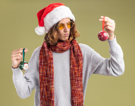 眼镜一个戴着圣诞老人帽 戴着黄色眼镜 脖子上围着暖和的围巾 手里拿着圣诞球的年轻人站在绿色的背景下 看起来很困惑 试图做出选择年轻温暖围着