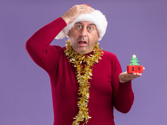 帽子中年男子戴着圣诞老人的帽子 脖子上戴着金属丝 手里拿着玩具方块 手里拿着25岁的约会对象 看着相机 心烦意乱地站在紫色背景上 手放在头上二十圣诞快乐中间