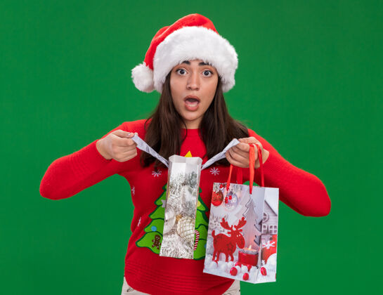女孩焦虑的年轻白人女孩戴着圣诞帽 手里拿着纸礼品袋 绿色背景上有复印空间礼物年轻帽子