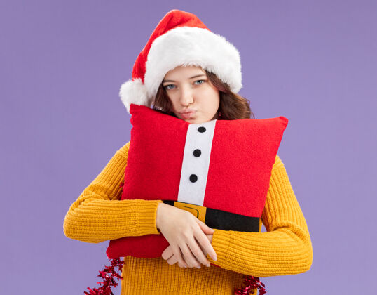 帽子自信的年轻斯拉夫女孩 戴着圣诞帽 脖子上戴着花环 抱着装饰好的枕头 看着紫色背景上的相机 还有复制空间拥抱圣诞快乐枕头