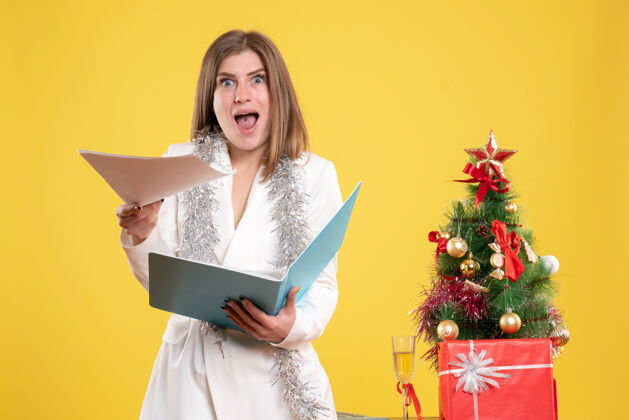 快乐正面图：女医生站着拿着文件放在黄色的桌子上 桌子上放着圣诞树和礼品盒肖像前面人